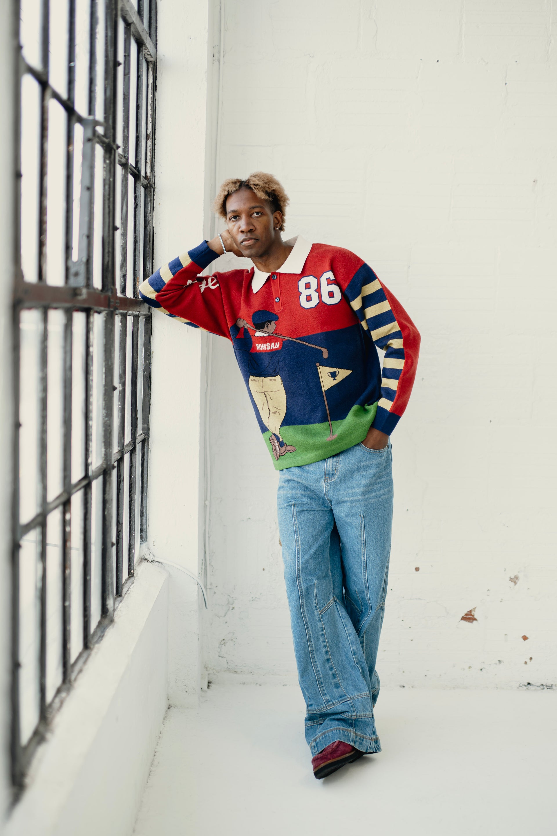Person wearing a colorful sweater and jeans leaning against a wall.